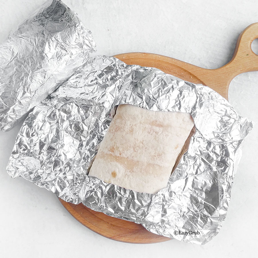 Wrapped burrito partially covered in aluminum foil on a cutting board.