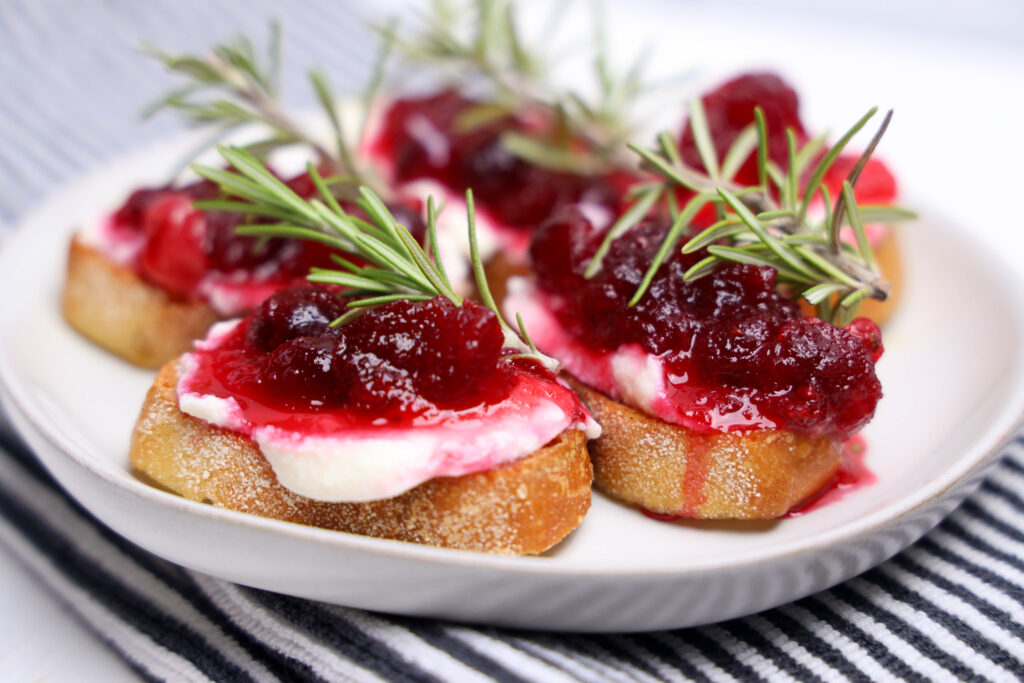Plate of small bread appetizers topped with white spread, red jam, and fresh herbs.