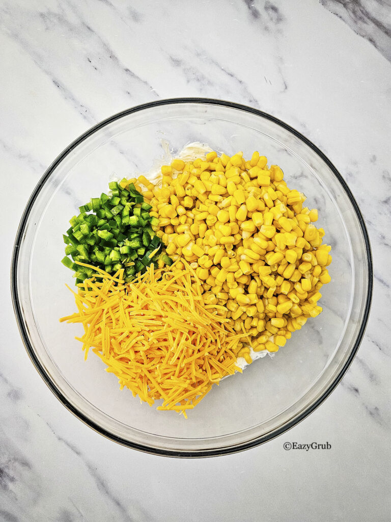 Unmixed corn kernels, diced peppers, and shredded cheese sitting over a cream base in a bowl.