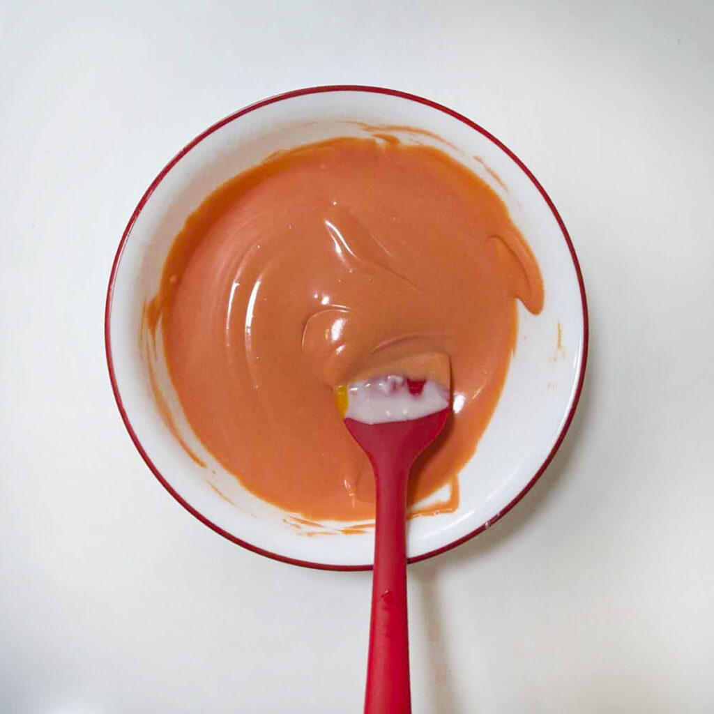 Orange tinted melted chocolate being stirred with a red spatula in a bowl